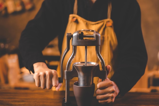 Barista sosteniendo un molinillo de mano para hacer bebidas moliendo granos de café tostados para una bebida de cafeína en la preparación del molino de desayuno matutino de fondo del café de la cocina con equipo de espresso en tono marrón