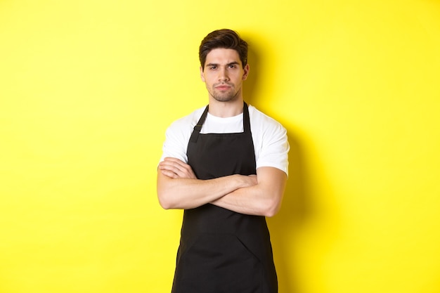 Barista suspeito apertando os olhos, cruzando os braços no peito e olhando para algo com descrença, de pé sobre um fundo amarelo