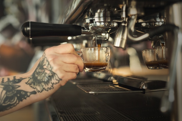 Barista tatuado fazendo espresso em uma moderna cafeteira. Close-up da mão feminina preparando uma bebida estimulante. Conceito de fabricação de café para pequenas empresas e profissionais