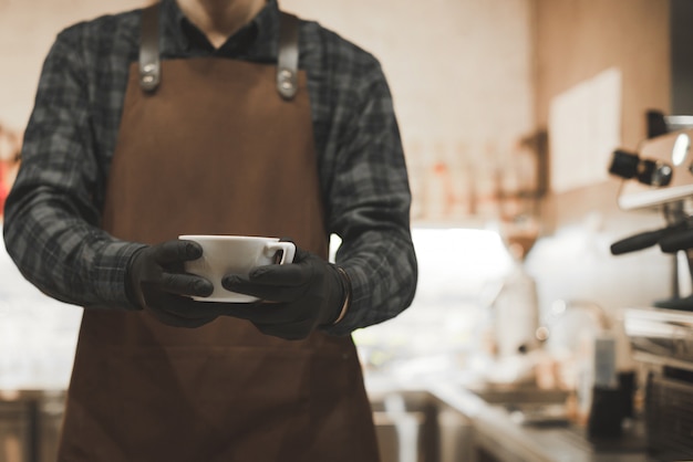 Baristas Mann in einer Schürze steht in einem gemütlichen Café in der Nähe einer Kaffeemaschine und hält eine Tasse Kaffee.