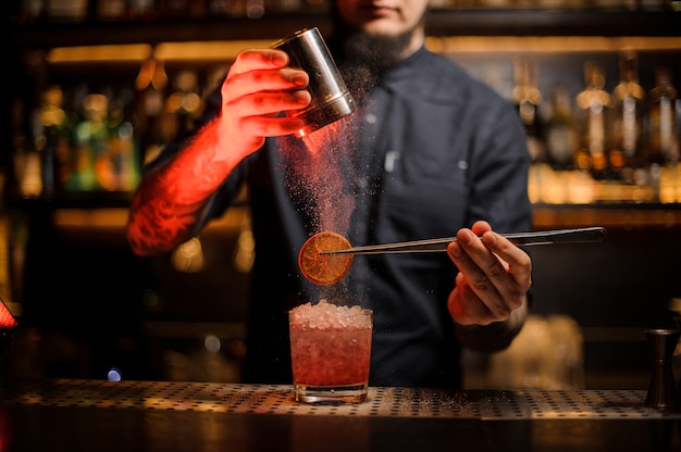 Foto barkeeper, der einem drink im glas eine getrocknete orange mit pinzette und aromatischem pulver auf der bartheke hinzufügt