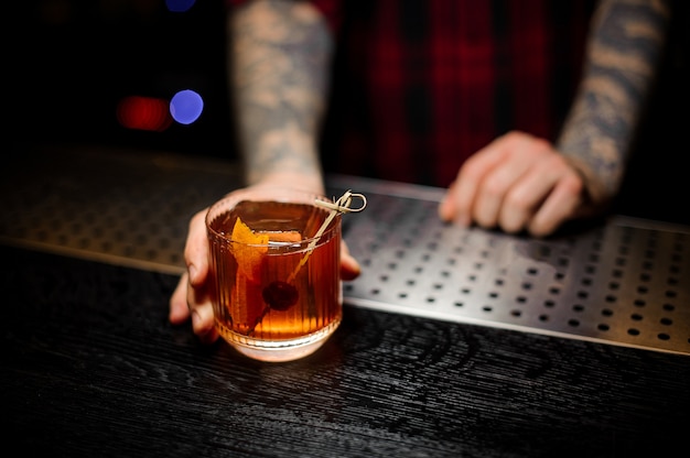 Barkeeper hält ein Glas Cocktail mit Whisky mit Orangenschale auf der Bartheke verziert