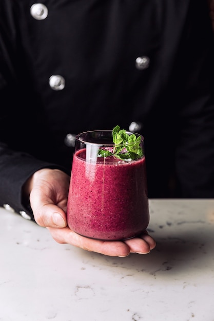 Barkeeper hält ein Glas erfrischenden Waldfruchtbeeren-Smoothie-Cocktail, garniert mit einem Minzzweig, Marmortisch