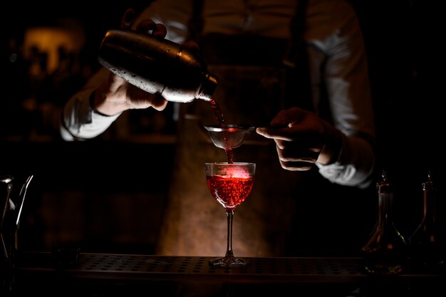 Barkeeper in Lederschürze gießt im Dunkeln ein rotes alkoholisches Getränk aus dem Stahlrüttler durch das Sieb