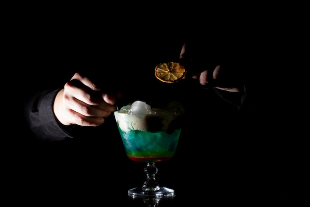 Barkeeper macht einen bunten Cocktail mit Eis und Schaum in einem Glas auf schwarzem Hintergrund
