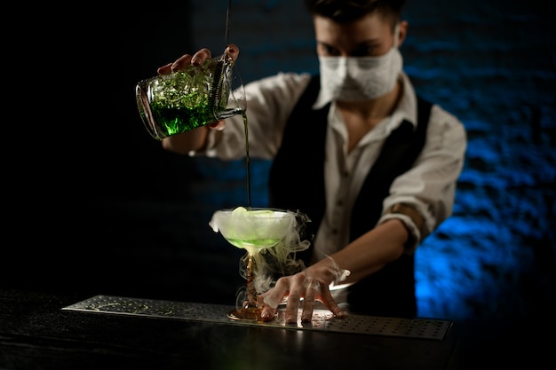 Barkeeper mit weißer medizinischer Maske gießt Cocktail in Glas