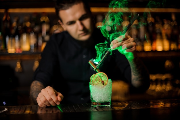 Foto barman adicionando ao copo uma laranja seca e canela defumada aromática gelada com uma pinça no balcão do bar