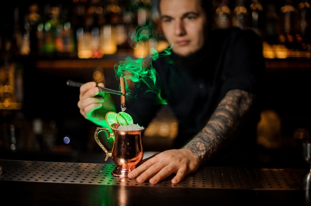 Barman añadiendo a un cóctel en el vaso de cobre con una canela ahumada aromática de naranja seca con pinzas en la barra del bar