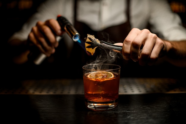 barman derritiendo caramelo con un quemador sobre el vaso de cóctel con una rodaja de limón en la oscuridad