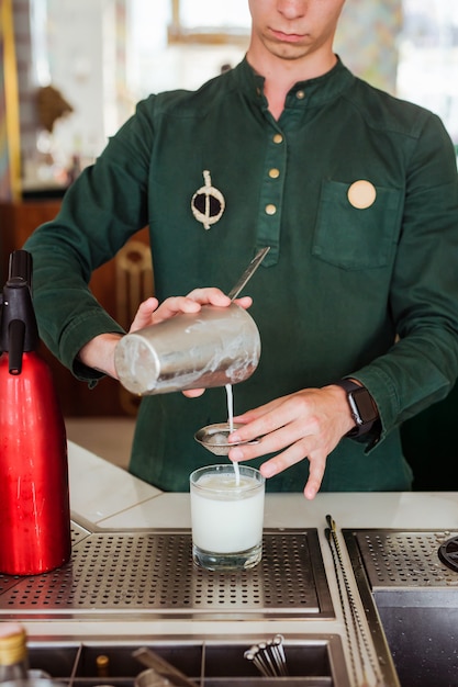 Barman fazendo um coquetel no bar: derramando uma bebida de uma coqueteleira através de uma peneira em um copo de vidro