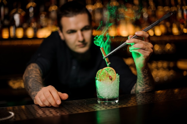 Barman masculino adicionando ao copo uma laranja seca e gelo picado e canela defumada aromática com uma pinça no balcão do bar