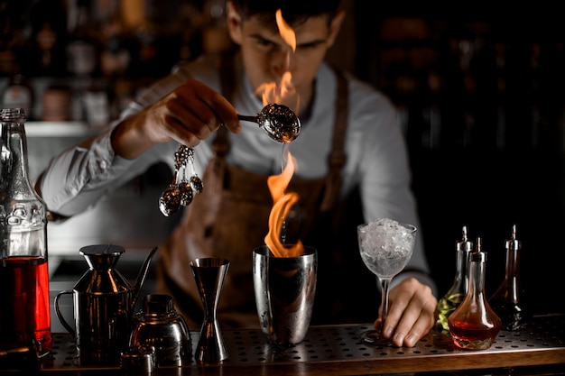 Barman masculino atraente, derramando uma essência da colher na chama para o agitador de aço