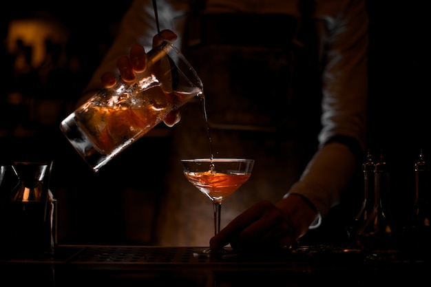 Foto barman masculino, derramando um coquetel alcoólico marrom do copo medidor para o copo no escuro