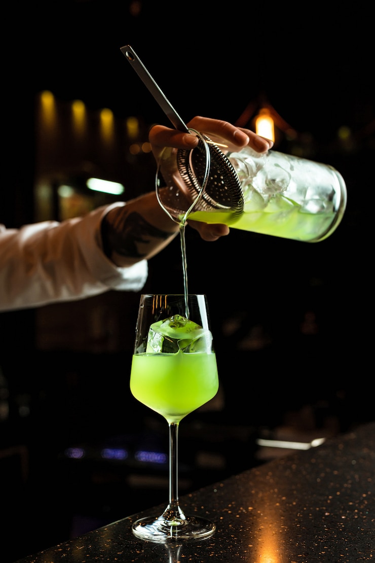 Foto barman masculino que prepairing um cocktail, derramando uma bebida verde de um copo de mistura através de um filtro em um copo de vinho com um cubo de gelo