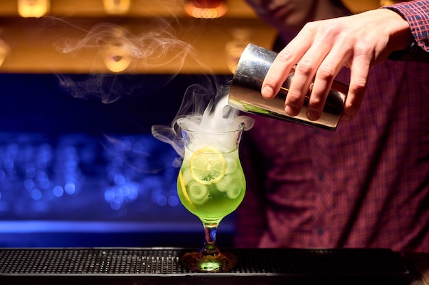 Barman preparando um coquetel alcoólico verde com rodelas de limão e gelo.