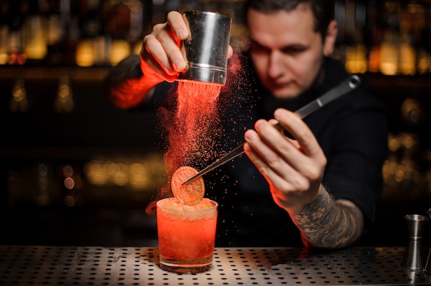 Barman profissional adicionando a um coquetel alcoólico no copo uma laranja desidratada com pinça e pó aromático no balcão do bar