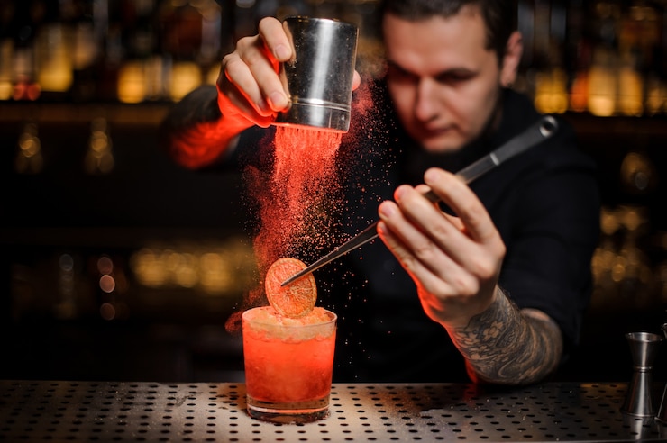 Foto barman profissional adicionando a um coquetel alcoólico no copo uma laranja desidratada com pinça e pó aromático no balcão do bar
