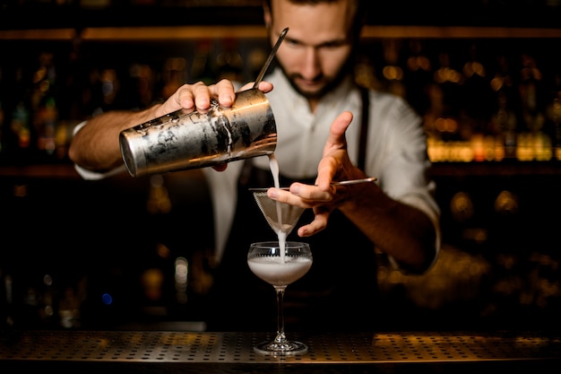 Foto barman profissional, derramando um coquetel branco da coqueteleira de aço no copo através da peneira