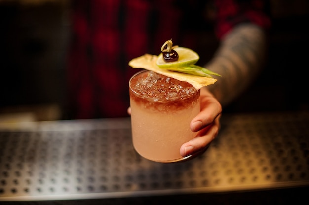 Barman segurando um copo de coquetel com uma saborosa bebida alcoólica doce com gelo e fatias de frutas no balcão do bar