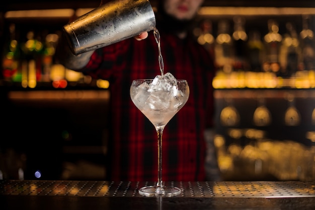 Barman servindo bebida alcoólica fresca do shaker em um copo com cubos de gelo contra as luzes