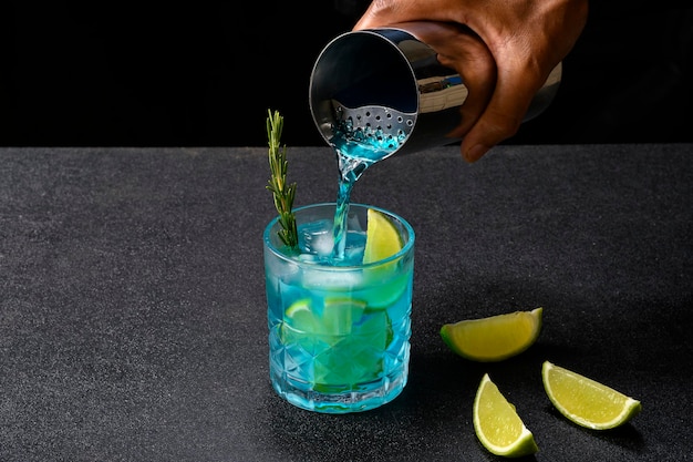 Barman servindo com uma coqueteleira uma bebida azul em um copo decorado com alecrim, gelo e limão preparando um coquetel, em fundo escuro.
