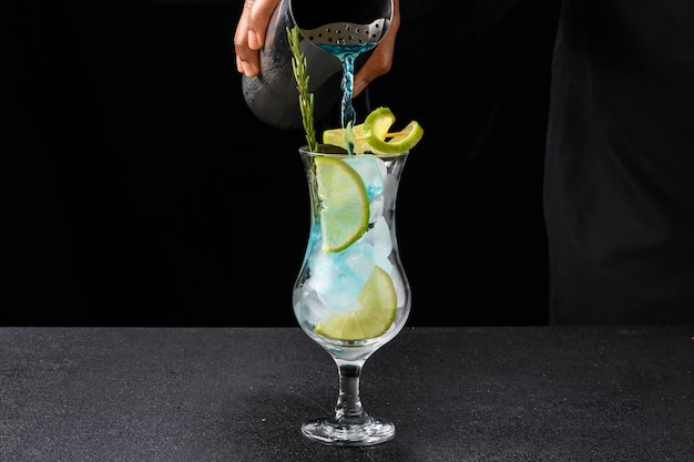 Barman servindo com uma coqueteleira uma bebida azul em um copo decorado com alecrim, gelo e limão preparando um coquetel, em fundo escuro.