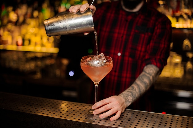 Barman servindo coquetel alcoólico em um copo cheio de cubos de gelo no balcão do bar