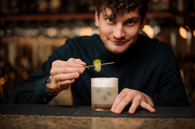 Barman sorridente, servindo um cocktail com um pepino no balcão de bar