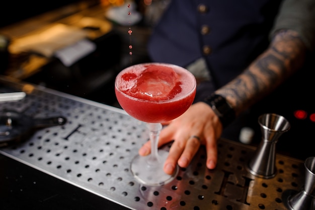 Barman con un tatuaje prepara un cóctel alcohólico rojo con hielo