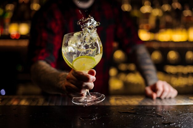 Barman con tatuajes sosteniendo una copa de cóctel con bebida cítrica dulce y agria fresca con rodajas de limón