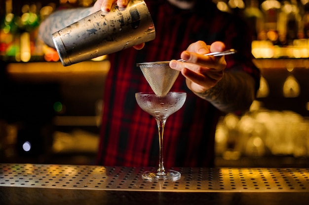 Barman vertiendo bebida alcohólica fresca en un vaso elegante con un colador en la barra