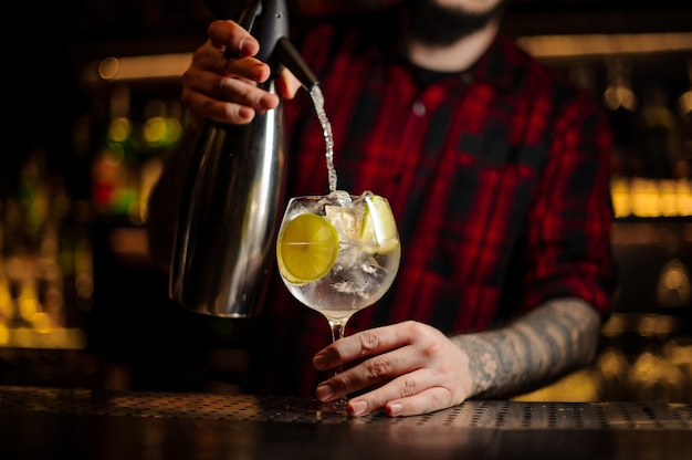 Barman vertiendo refrescos en una copa de cóctel con bebida alcohólica y limón en la barra del bar