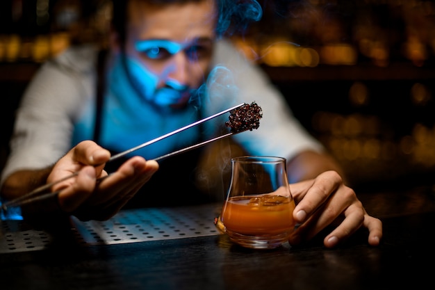 Barmixer, der gekühltes schmelzendes Karamell mit Twezzers dem Cocktail mit Eiswürfeln unter Blaulicht hinzufügt