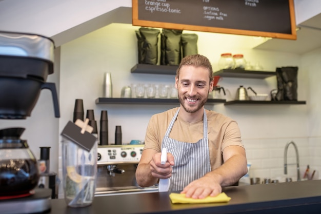 Barra de bar, limpieza. Alegre joven adulto de pie detrás de la barra haciendo limpieza en húmedo limpiando superficies con una servilleta