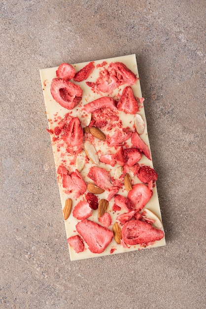 Una barra de chocolate blanco con almendras fritas y fresas liofilizadas sobre un fondo claro. Vista superior, endecha plana.