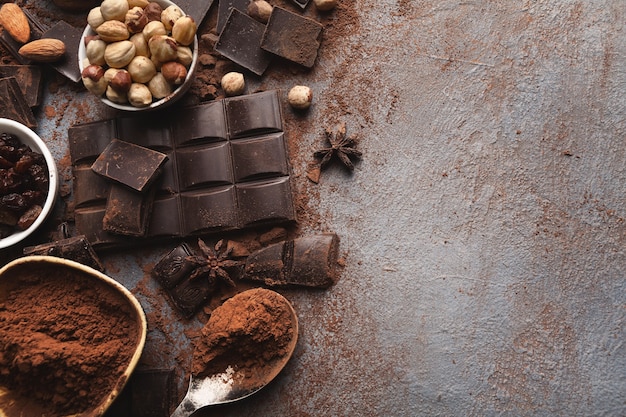 Barra de chocolate escuro quebrada em pedaços em fundo escuro, cacau em pó e várias nozes em tigelas closeup, copie o espaço. papel de parede doce, publicidade de confeitaria e conceito de ingredientes para cozinhar