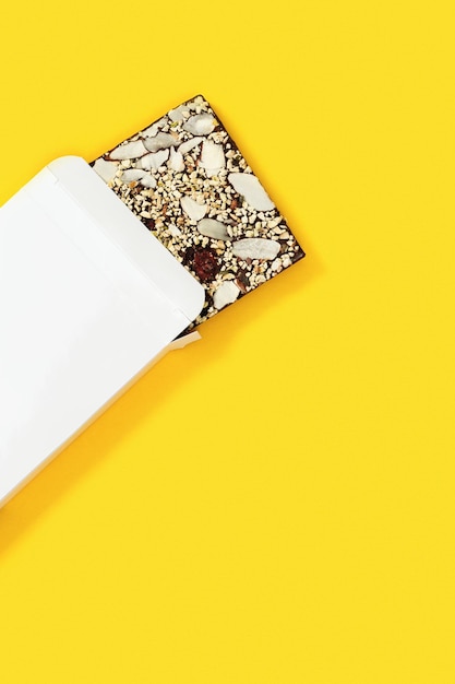 Barra de chocolate mordida com nozes e frutas secas em embalagem de papel branco sobre fundo amarelo