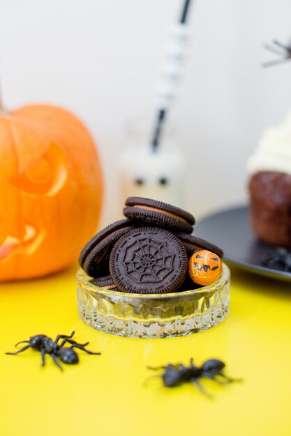 Barra de dulces de comida de Halloween con dulces y calabaza tallada en una mesa amarilla Galletas negras con telaraña grandes hormigas negras y pastelitos con crema