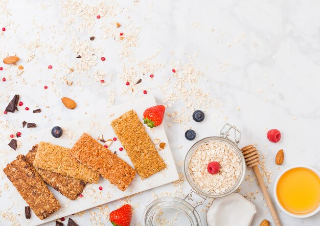 Barra de granola de cereales orgánicos con bayas en tablero de mármol con cuchara de miel y tarro de avena y coco en mesa de mármol.