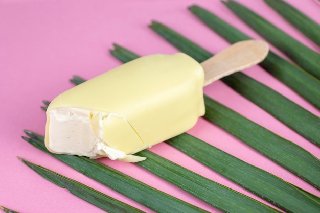 Barra de helado de vainilla en palo recubierto con chocolate blanco sobre fondo rosa con una hoja de palma