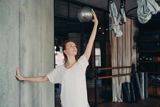 Barré-Training. Lächelnde Frau in Sportkleidung, die in der Hand über ihrem Kopf zu einem silbernen Pilates-Ball blickt und mit einer anderen Hand die Wand berührt, während sie in einem modernen Fitnessstudio trainiert