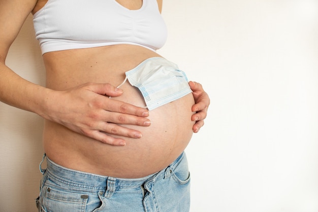 Barriga de grávida com máscara protetora médica sobre fundo claro. Gravidez conceitual em epidemia de coronavírus, problemas de gravidez, intoxicação, aborto, risco de aborto espontâneo, pandemia de covid-19, doença