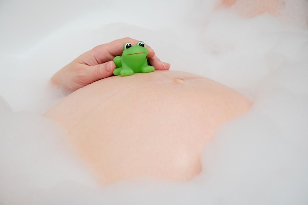 Barriga de gravidez com brinquedo de sapo verde closeup Mulher grávida tomando banho de espuma