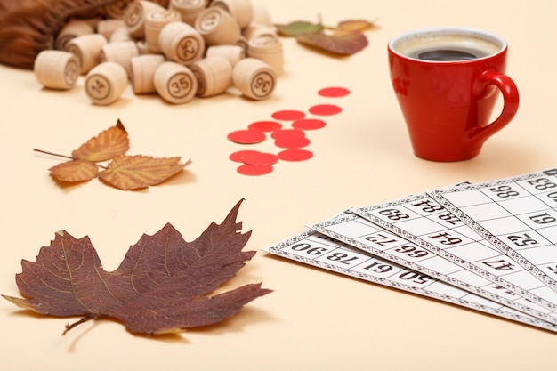 Foto barriles de lotería de madera con bolsa abierta, hojas secas de otoño, taza de café y cartas de juego sobre fondo beige. juego de mesa de lotería. tema de otoño.