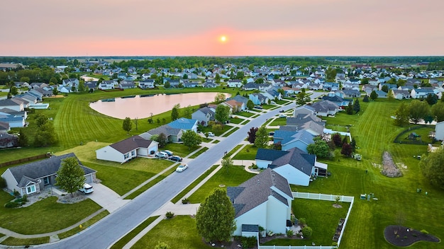 Barrio suburbano al atardecer con estanque, casas de uno y dos pisos, césped verde, antena de sol brillante de color naranja