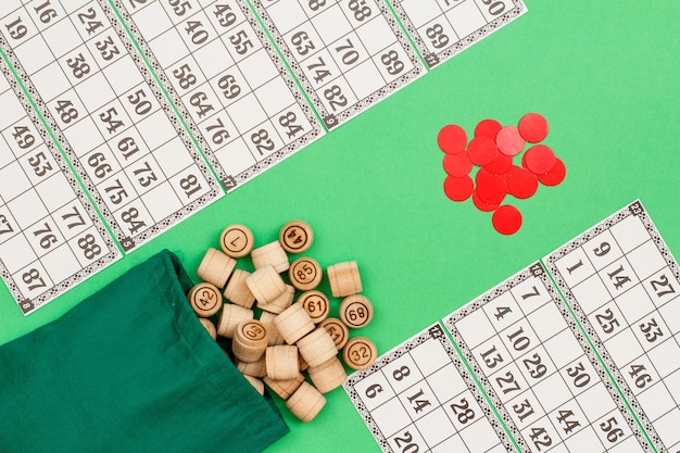 Foto barris de loto de madeira com saco de pano, cartas de jogos e fichas vermelhas sobre fundo verde.