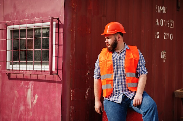 Bartarbeitskraftmann-Klagenbauarbeiter im orange Sturzhelm der Sicherheit, der auf rotem Fass sitzt.