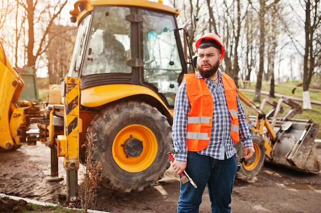 Bartarbeitskraftmann-Klagenbauarbeiter im orange Sturzhelm der Sicherheit, gegen Traktor mit dem justierbaren Schlüssel zur Hand.