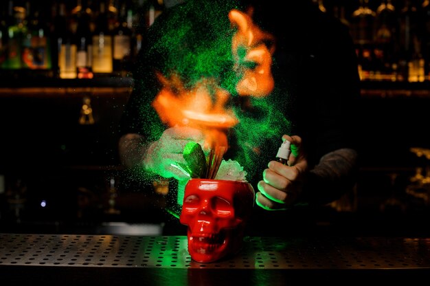 Bartender profesional rociando el cóctel en la taza de scull roja del vaporizador y flameándolo con la luz verde en la barra del bar.