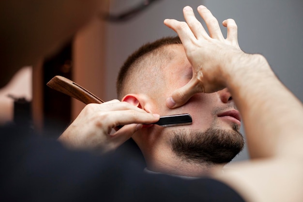 Barthaarschnitt in einem Friseursalon, ein junger Mann, der seinen Bart mit einem Rasiermesser in der Nähe rasiert